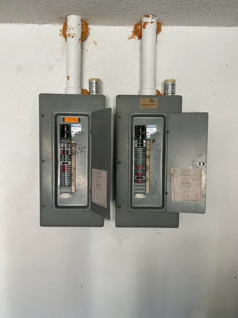 Two gray electrical panels with breaker boxes and white PVC conduit risers mounted on wall